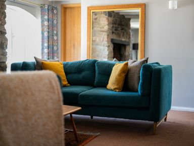 A living room with a teal sofa and yellow and brown pillows with a large mirror reflecting a stone wall at The Arches in Bamburgh