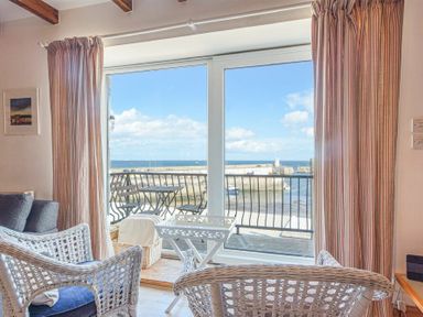 A seating area with wicker chairs and a small white table facing a balcony with a view of the harbor and sea at Farne Lookout in Seahouses