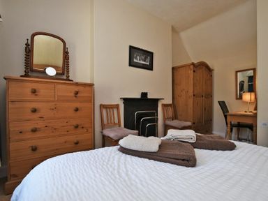 A bedroom with a wooden chest of drawers mirror bed with towels chairs wardrobe and desk with lamp at Turnstone Cottage Nr Bamburgh