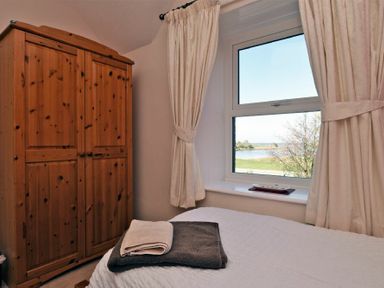A bedroom with a wooden wardrobe and a bed near a window with curtains showing a view of water and trees at Turnstone Cottage Nr Bamburgh