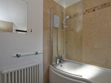A bathroom with a bathtub shower unit glass shelf radiator and tiled walls at Turnstone Cottage Nr Bamburgh