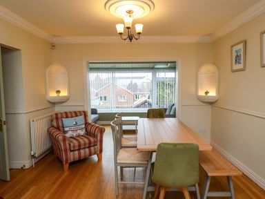 A dining room with a wooden table green and beige chairs a striped armchair and a view of a sunroom with windows at Barnacle Cove in Beadnell