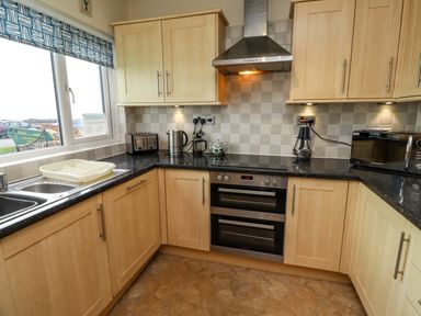 A kitchen with wooden cabinets a black countertop a double stainless steel sink a toaster kettle coffee press microwave and oven at Barnacle Cove Beadnell