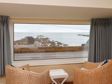 A room with two wicker chairs and a small white table by a large window overlooking a rocky shoreline and sea at Barnacle Cove in Beadnell
