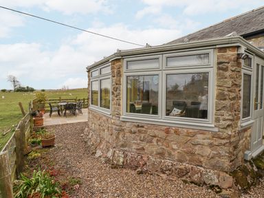 A stone exterior of a house with large windows and a patio table and chairs in a field at Adderstone Cottage in Bamburgh
