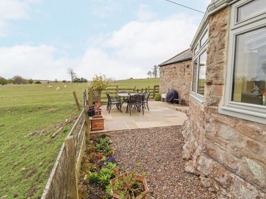 An outdoor patio with a metal table and chairs next to a stone house and a fenced grassy field with sheep at Adderstone Cottage in Bamburgh