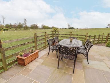 An outdoor patio with a metal table and six chairs overlooking a fenced grassy field with sheep at Adderstone Cottage in Bamburgh