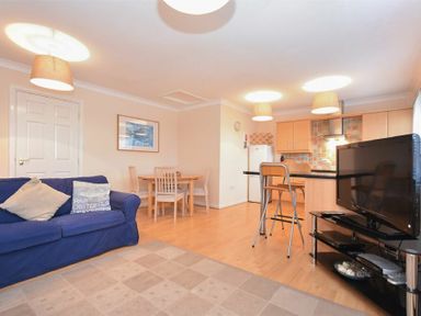An open-plan living area with a blue sofa a dining table with chairs and a kitchen with a breakfast bar and stools at Beachcomber Apartment in Bamburgh