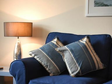 A blue sofa with two striped pillows and a table lamp on a side table at Beachcomber Apartment in Bamburgh