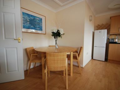 A dining area with a round wooden table and four wicker chairs a vase with flowers on the table a refrigerator and kitchen cabinets in the corner at Beachcomber Apartment in Bamburgh
