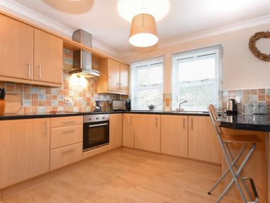 A kitchen with wooden cabinets black countertops tiled backsplash and two windows at Beachcomber Apartment in Bamburgh