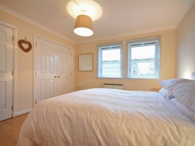 A bedroom with a double bed, two windows with blinds, a ceiling light, a wall mirror, a heart-shaped wall decoration, and two closed white doors at Beachcomber Apartment in Bamburgh
