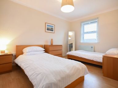 A bedroom with two single beds a bedside table a chest of drawers a standing mirror and a window at Beachcomber Apartment in Bamburgh