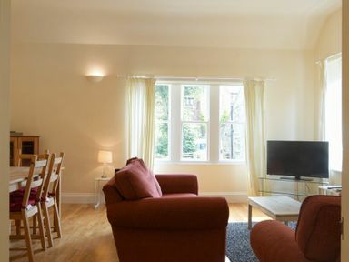 A living room with red sofas a wooden dining table and chairs a TV on a glass stand and large windows with curtains at Decked Out in Alnmouth