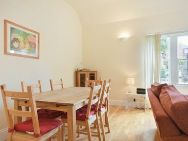 A dining area with a wooden table and six chairs with red cushions next to a brown sofa and a small table with a lamp and radio by the window at Decked Out in Alnmouth