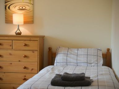 A bedroom with a wooden bed and dresser with a lamp and folded towels at Decked Out in Alnmouth