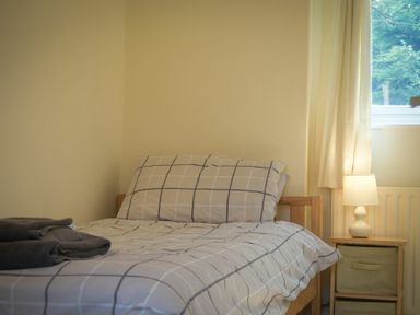 A bedroom with a single bed with checkered bedding a bedside table with a lamp and folded towels near a window at Decked Out in Alnmouth