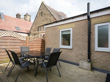 An outdoor patio area with a glass table and six black chairs adjacent to a brick building with two windows surrounded by wooden fencing at Decked Out in Alnmouth