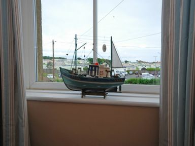 A model fishing boat on a windowsill with a view of cars and houses outside at Coastal Haven in Seahouses
