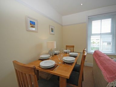 A dining area with a wooden table set for six with plates glasses and cutlery next to a window with blinds and a sofa with a red throw at Coastal Haven in Seahouses