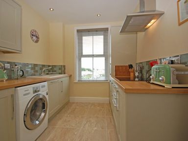 A kitchen with cabinets a washing machine a sink a window and a countertop with a toaster and utensil holder at Coastal Haven in Seahouses