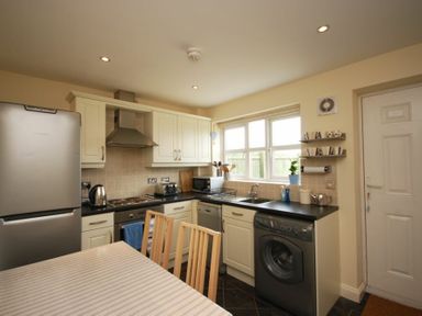 A kitchen with white cabinets black countertops a silver refrigerator a washing machine a dining table with wooden chairs and a window at House Martins Beadnell