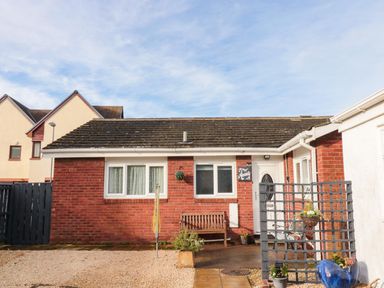 A small single-story brick house with a white door windows a wooden bench a hanging plant a garden trellis and a blue ceramic pot at Arcadie in Amble