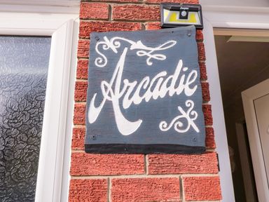 A black wooden sign with white lettering that reads Arcadie mounted on a red brick wall near a window and a door at Arcadie in Amble