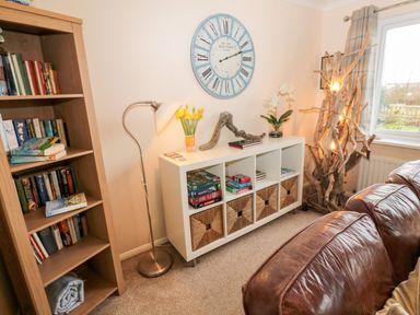 A living room with a bookshelf a floor lamp a white cubed storage unit with baskets books and decorations a clock on the wall and brown leather sofas at Arcadie in Amble