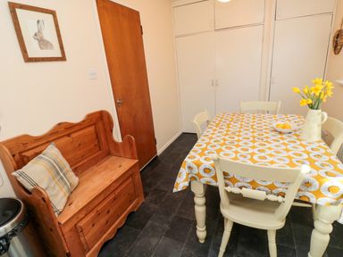 A kitchen with a wooden bench with a cushion a table with a patterned cloth and chairs white cabinets and a door at Arcadie in Amble