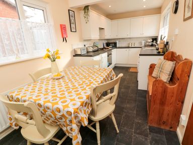 A kitchen with a dining table covered with a patterned tablecloth and chairs a wooden bench a countertop with kitchen appliances and cabinets at Arcadie in Amble