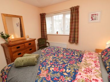 A bedroom with a bed covered in floral bedding and towels on top a wooden dresser with a mirror green chair and window with check curtains at Arcadie in Amble