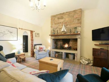 A living room with a stone fireplace a television mounted on the wall and upholstered chairs at Charlton Cottage in Bamburgh