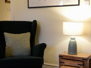 A black armchair with a beige cushion next to a wooden side table with a gray lamp on it at Charlton Cottage in Bamburgh