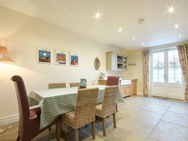 A dining area with a table covered by a tablecloth and wicker chairs next to a kitchen with cabinets and a sink at Charlton Cottage Bamburgh