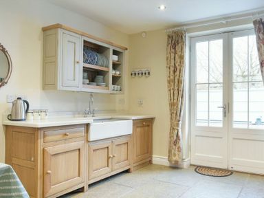 A kitchen with wooden cabinets a white farmhouse sink open shelves with plates a kettle and floral curtains by glass doors at Charlton Cottage in Bamburgh