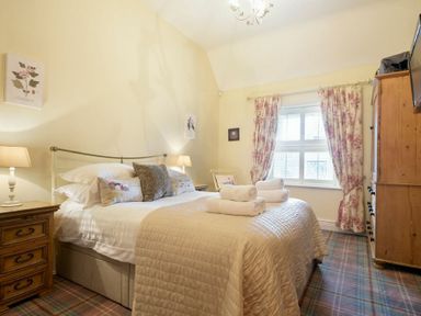 A bedroom with a bed a wooden nightstand lamps floral curtains and a wooden wardrobe at Charlton Cottage in Bamburgh