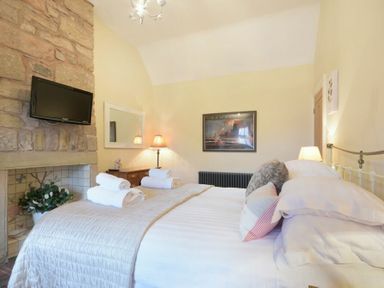 A bedroom with a bed and pillows a wall-mounted tv and a stone fireplace at Charlton Cottage in Bamburgh