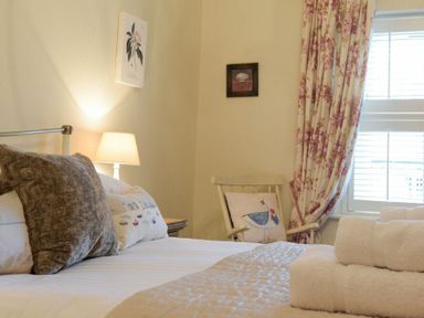 A bedroom with a bed with pillows a lamp on a bedside table a chair with a cushion floral curtains and rolled towels on the bed at Charlton Cottage in Bamburgh