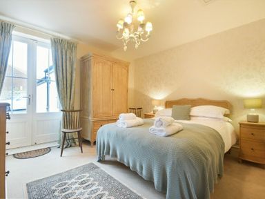 A bedroom with a bed covered with a blanket and towels two bedside tables lamps a wooden wardrobe chairs and a rug at Charlton Cottage in Bamburgh