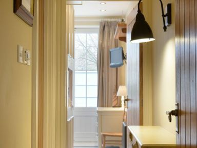 A narrow hallway with wooden doors wall lamp and a view of a room with a window television and a chair at Charlton Cottage in Bamburgh