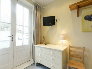 A room corner with double glass doors curtain white dresser wooden chair small lamp and wall mounted tv at Charlton Cottage Bamburgh