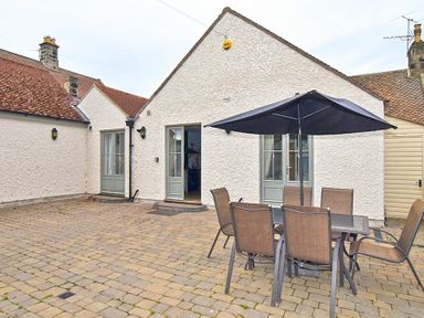 An outdoor patio area with a table eight chairs and a large umbrella in front of a white building with multiple doors at Charlton Cottage in Bamburgh