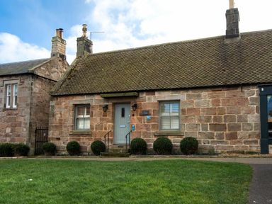 A stone cottage with a grey door and two windows with bushes in front at Charlton Cottage in Bamburgh