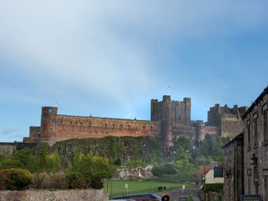 A large historic stone castle on a hill with greenery around and buildings along a street at Charlton Cottage in Bamburgh