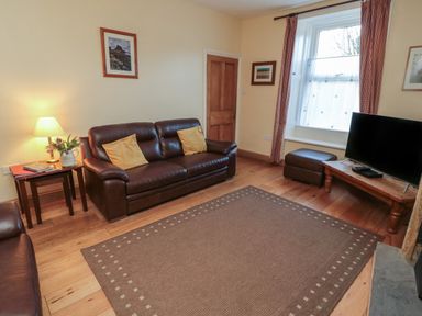 A living room with brown leather sofas a wooden coffee table a television a window with curtains and a lamp on a side table at Riverside Cottage in Warkworth