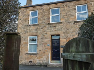 The stone exterior of a two-story house with white framed windows and a black door behind a wooden gate at Riverside Cottage in Warkworth