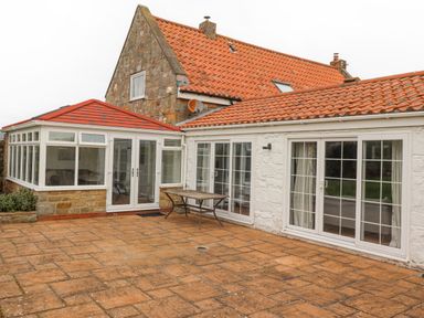 A stone house with red tiled roof and glass doors opening to a patio with a wooden table at Jara Bamburgh