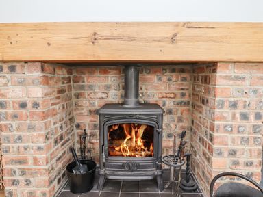 A wood burning stove with fire inside set in a brick fireplace with fire tools and bucket at Jara in Bamburgh