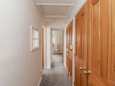 A hallway with grey carpet wooden doors on the right and a chair in the room at the end of the corridor at Jara in Bamburgh